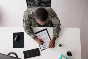 Aerial shot of a man in a U.S. military uniform sitting at a white desk, filling out paperwork.